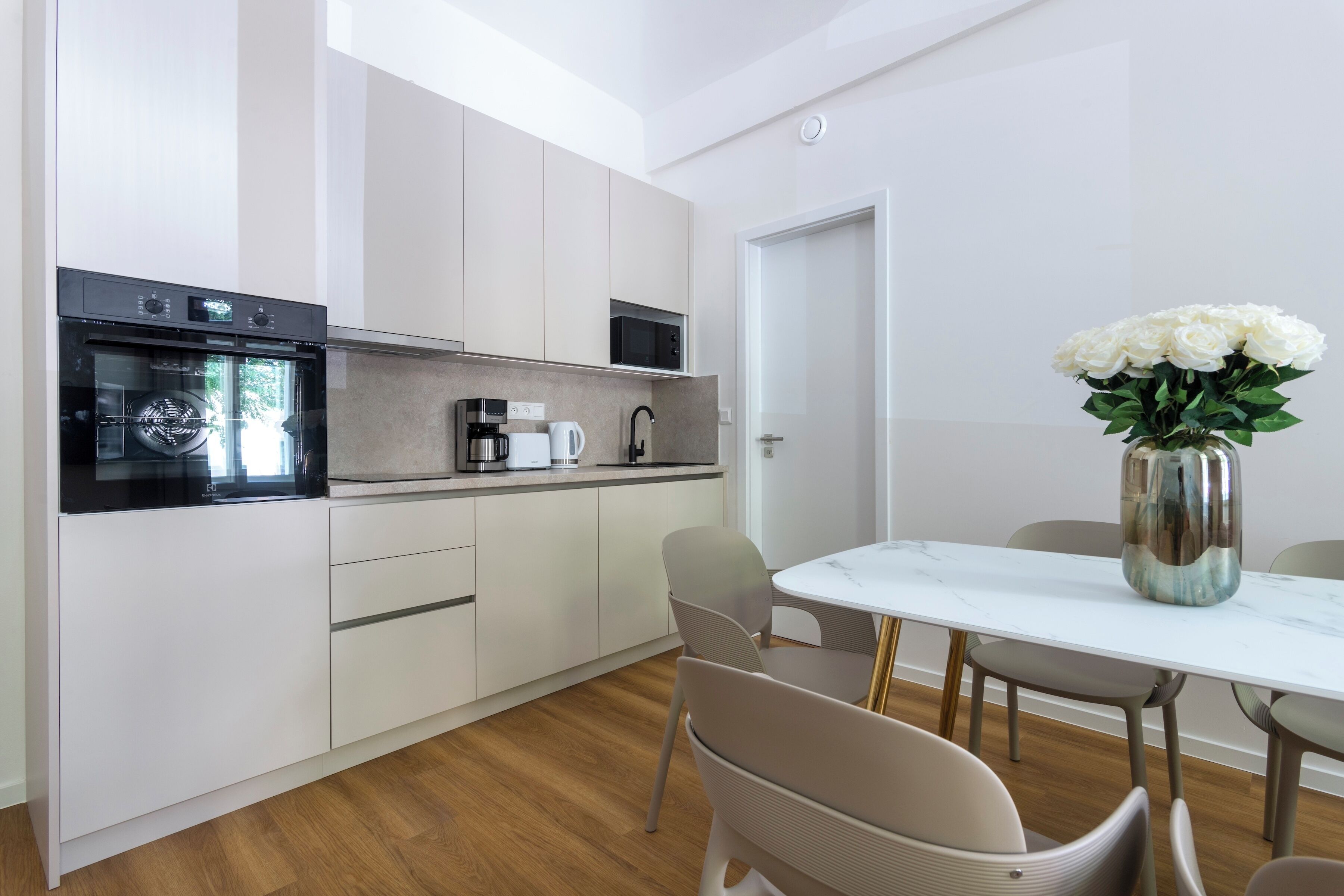 A modern kitchen with a built-in oven and microwave, a dining table with chairs, and a vase with white flowers.