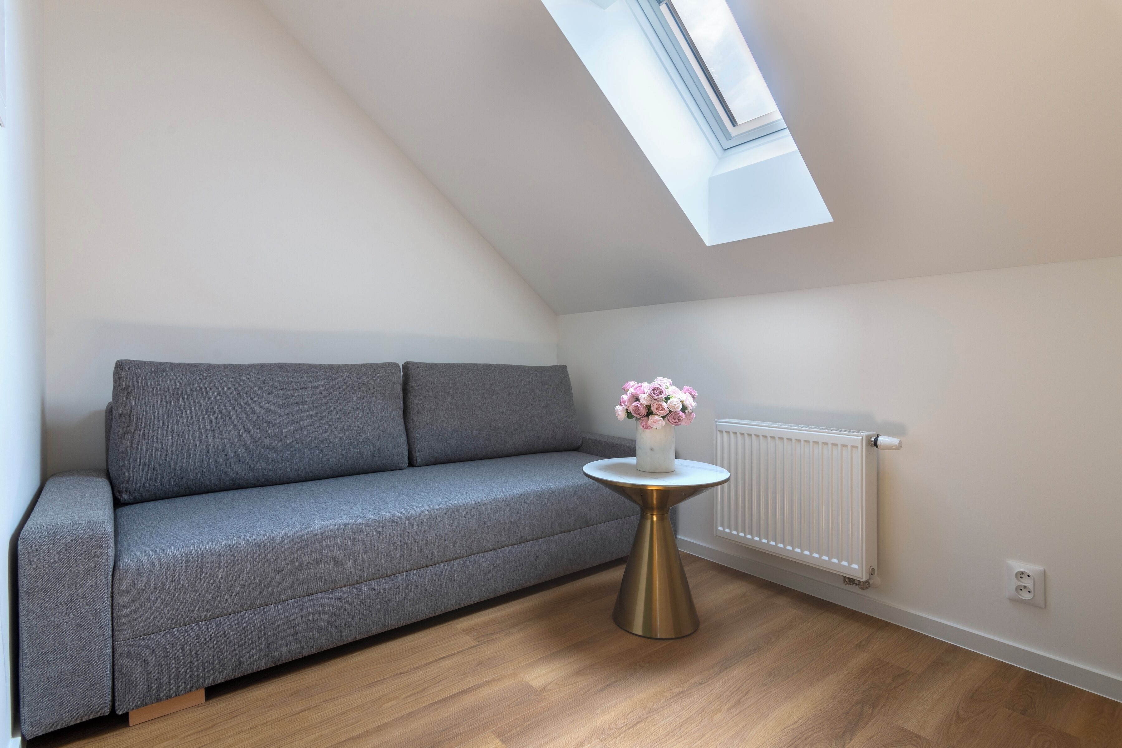 A modern living room with a grey sofa, a small round table with a vase of flowers, and a skylight above.