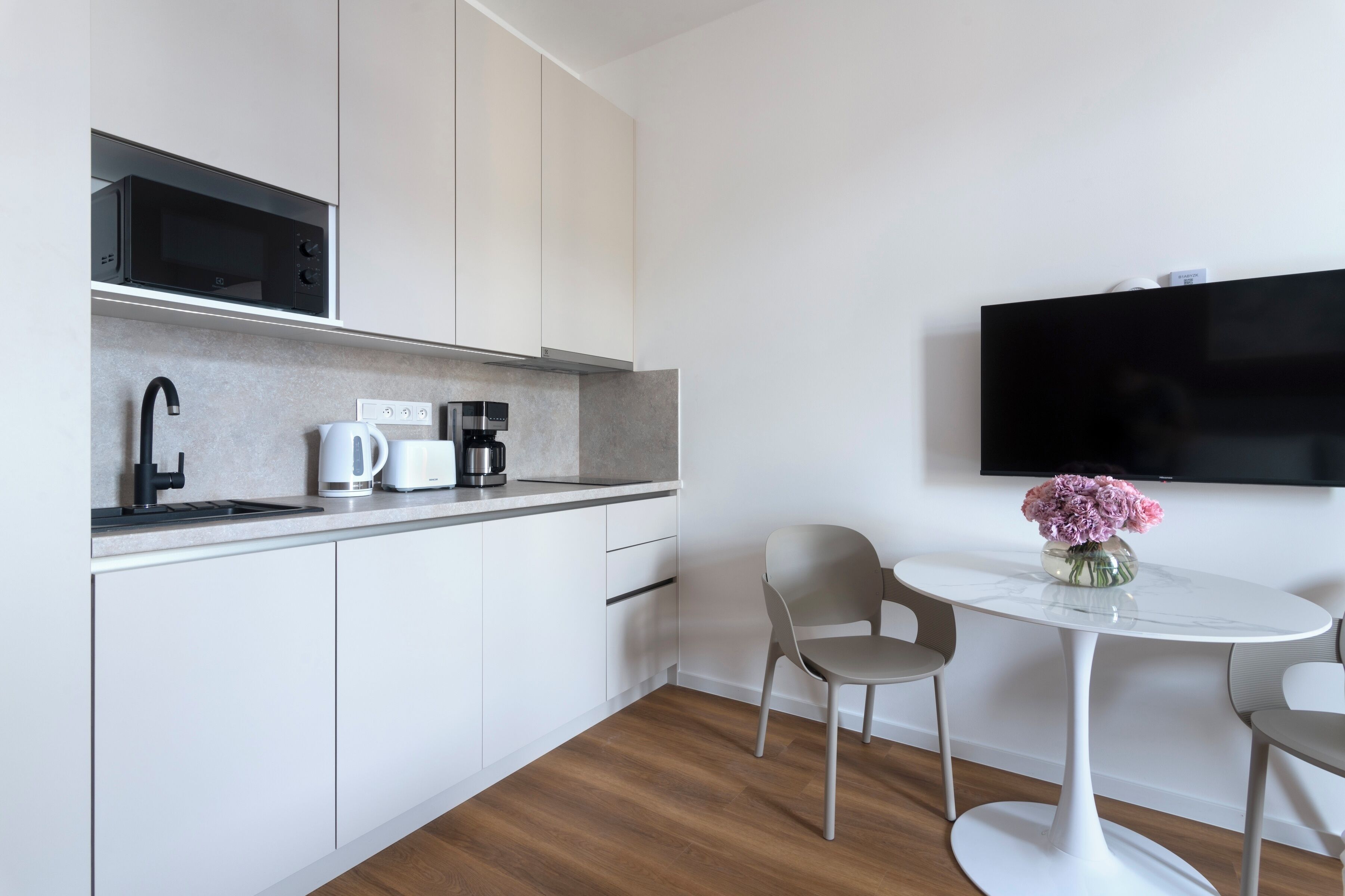 A modern kitchen with a microwave, coffee maker, and kettle, a small dining table with chairs, and a flat-screen TV mounted on the wall.