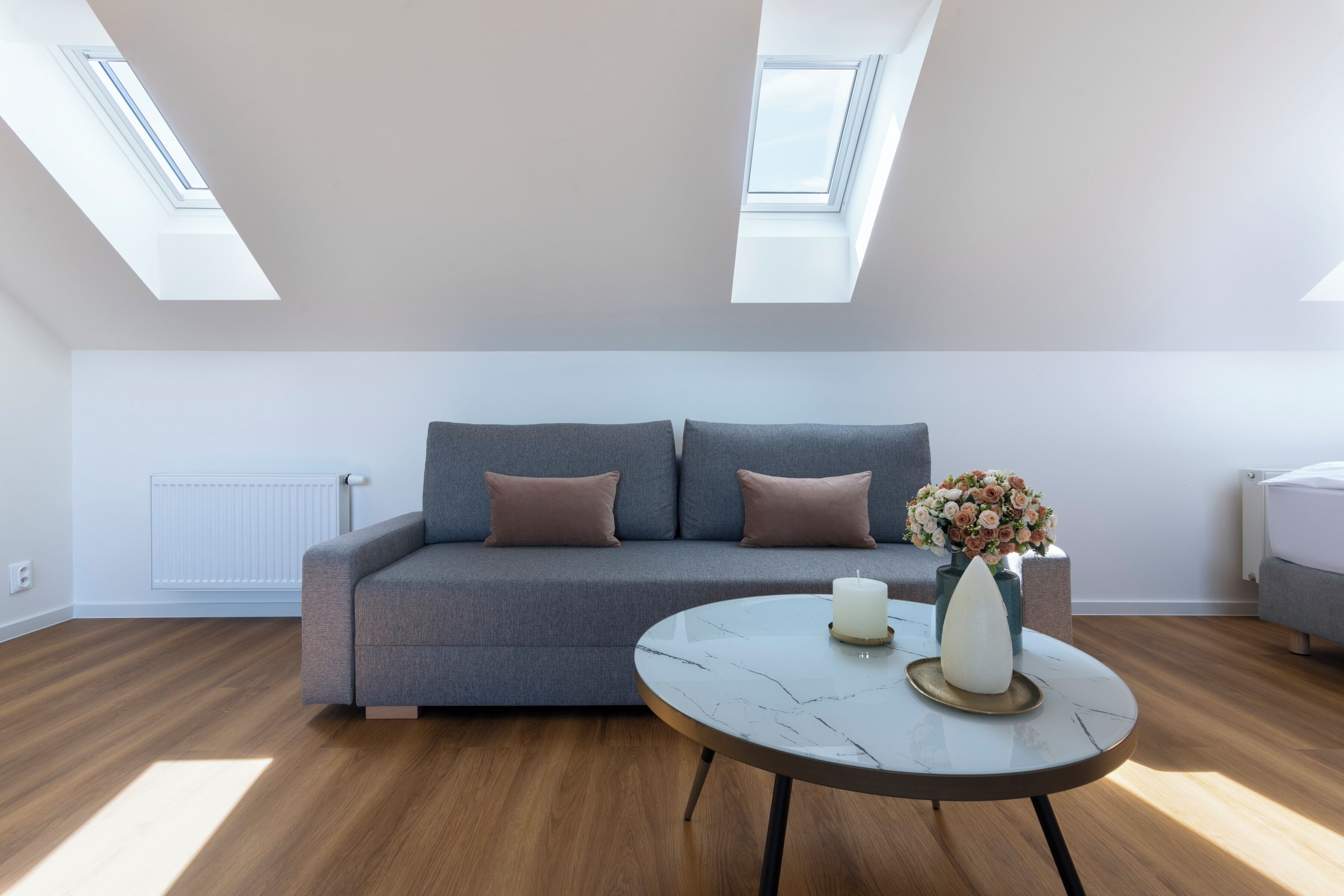 Una sala de estar moderna con un sofá gris, una mesa redonda de mármol y un jarrón con flores. Hay tragaluces sobre el sofá.