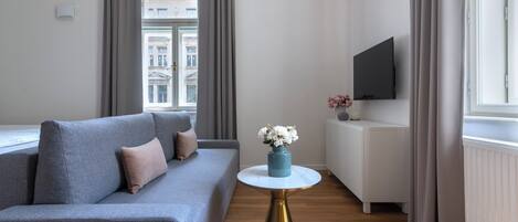 Un soggiorno moderno con un divano grigio, un tavolino rotondo con un vaso di fiori, un televisore a schermo piatto su un mobile bianco e una finestra con tende.