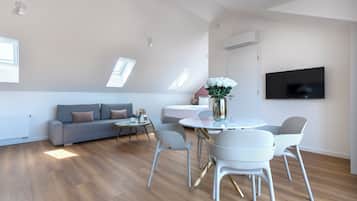 A modern living room with a sofa, a dining table with chairs, a flat-screen TV mounted on the wall, and a skylight.