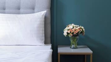 En pænt redt seng med hvide sengetæpper, en grå polstret sengegavl og et natbord med en vase med blomster.