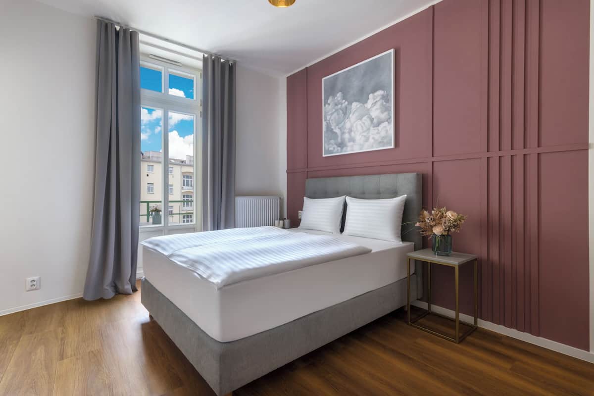 Ein modernes Schlafzimmer mit einem großen Bett, Blick auf Gebäude durch das Fenster und einem gerahmten Kunstwerk an der Wand.