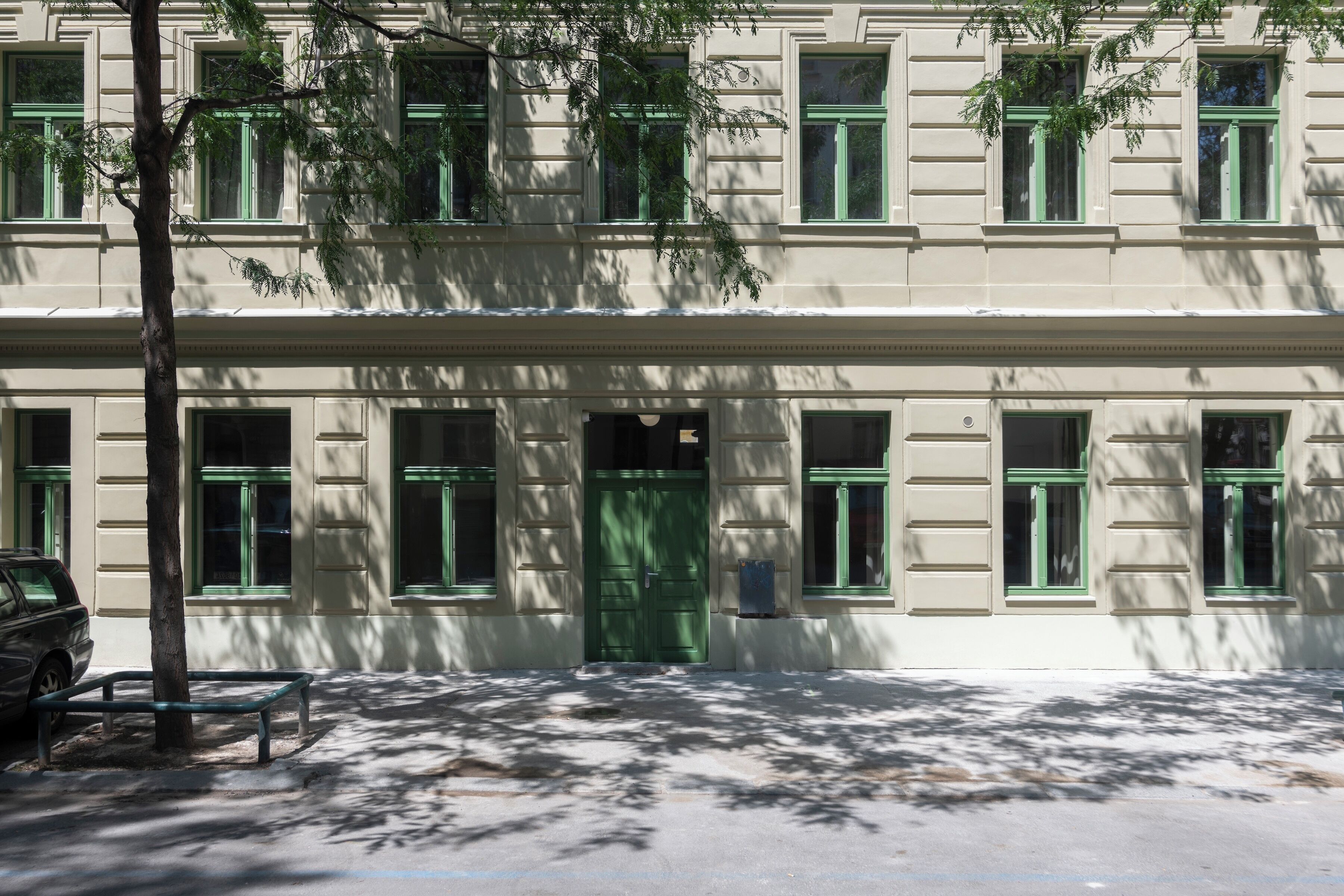 A building with a green door and multiple windows, a bench, and a tree in front.