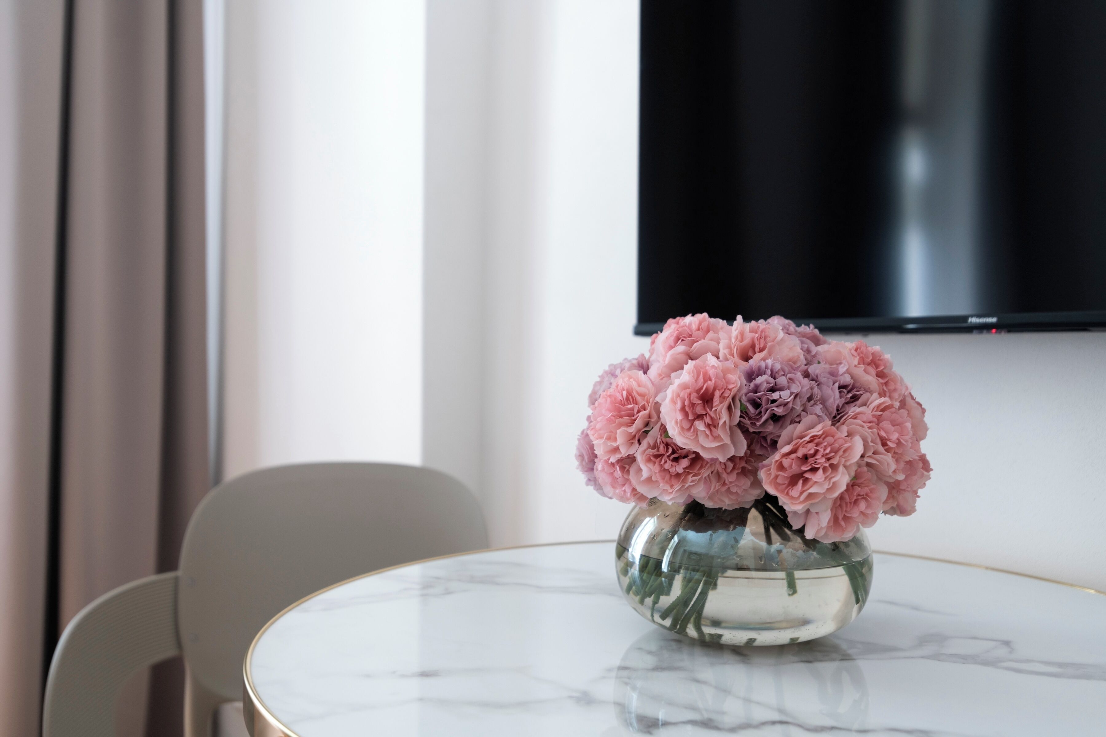 Una mesa de mármol con un jarrón de flores rosadas, una silla moderna y un televisor de pantalla plana montado en la pared.