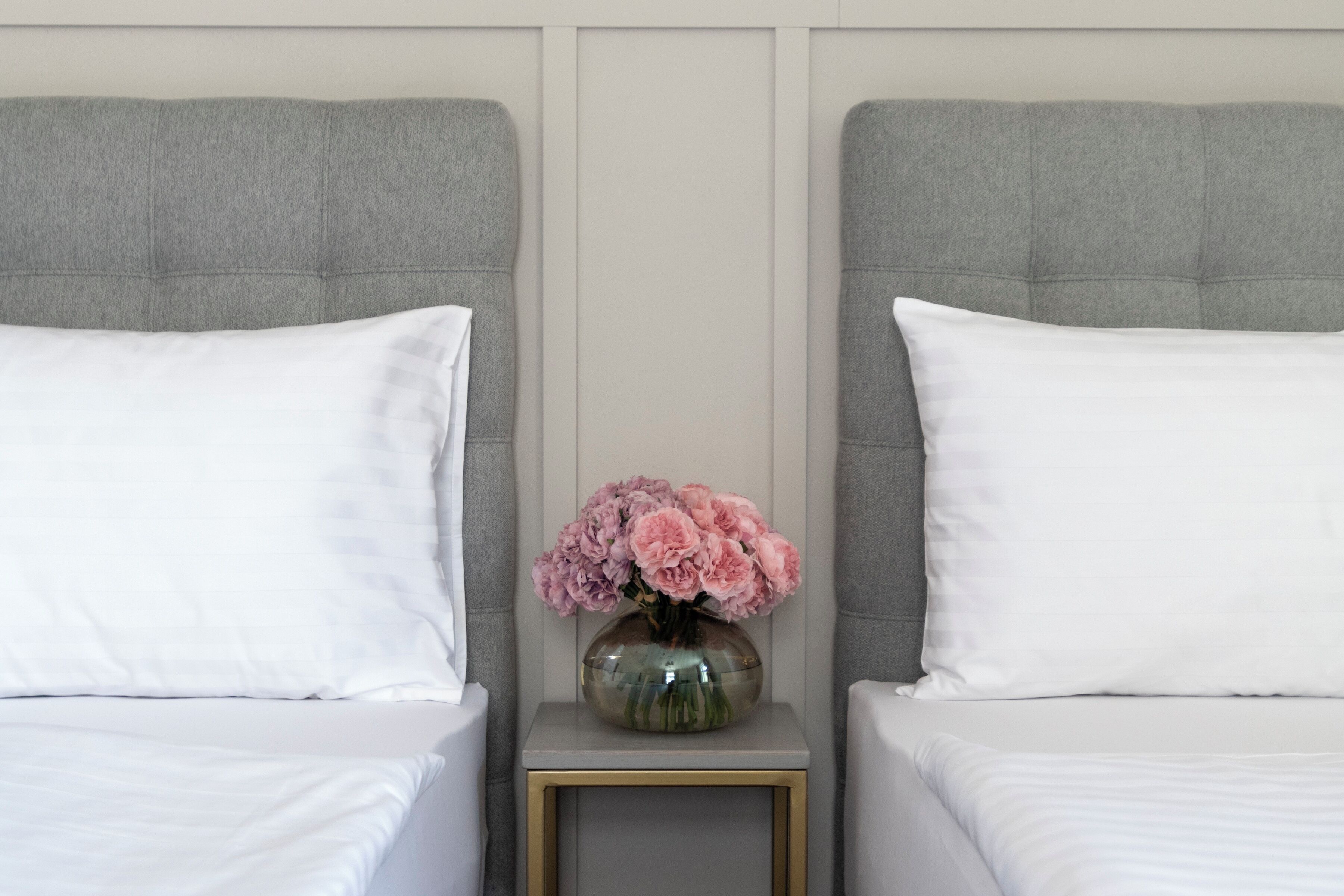 Ein Hotelzimmer mit zwei Betten, jeweils mit weißer Bettwäsche und grauem Kopfteil. Ein kleiner Tisch zwischen den Betten trägt eine Vase mit rosa Blumen.