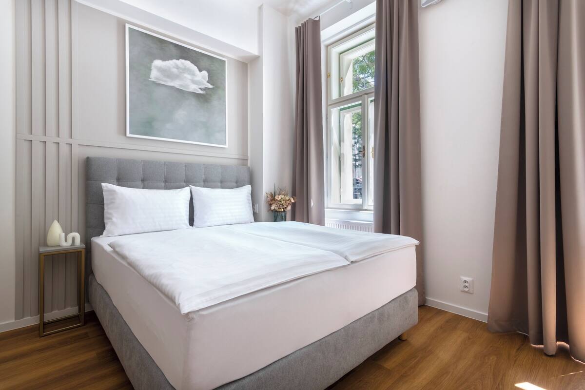 Ein ordentlich bezogenes Bett mit weißen Leinentüchern in einem modernen Schlafzimmer mit grauem Kopfteil, einem gerahmten Kunstwerk an der Wand und einem Fenster mit Vorhängen.