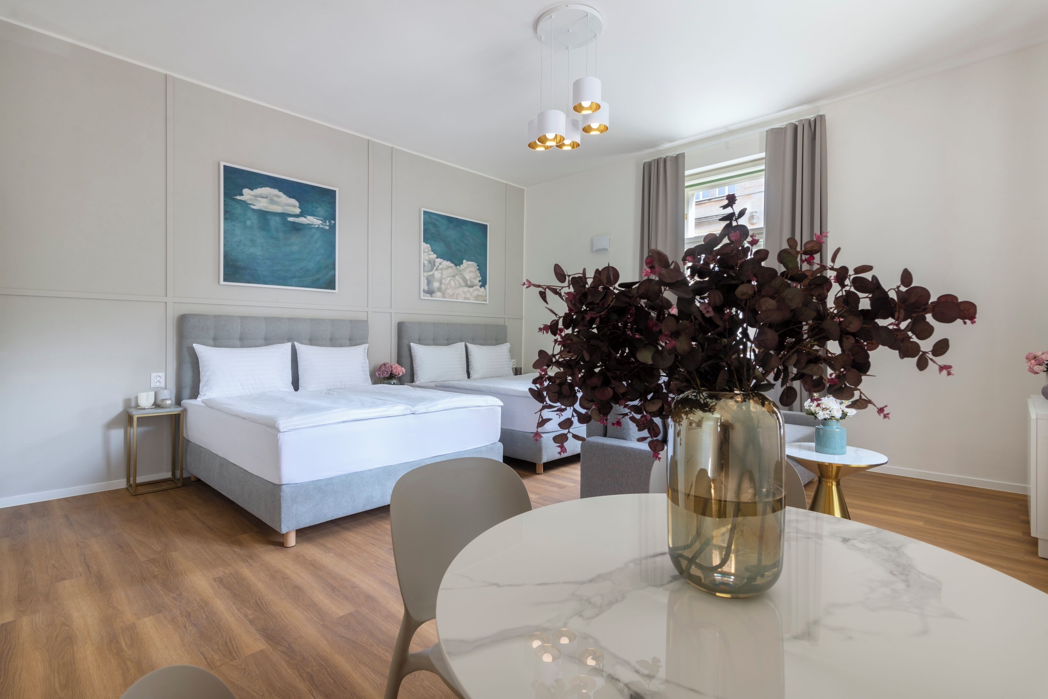 Moderni hotellihuone, jossa on sänky, pyöreä ruokapöytä, maljakko kukkineen ja taideteoksia seinillä.