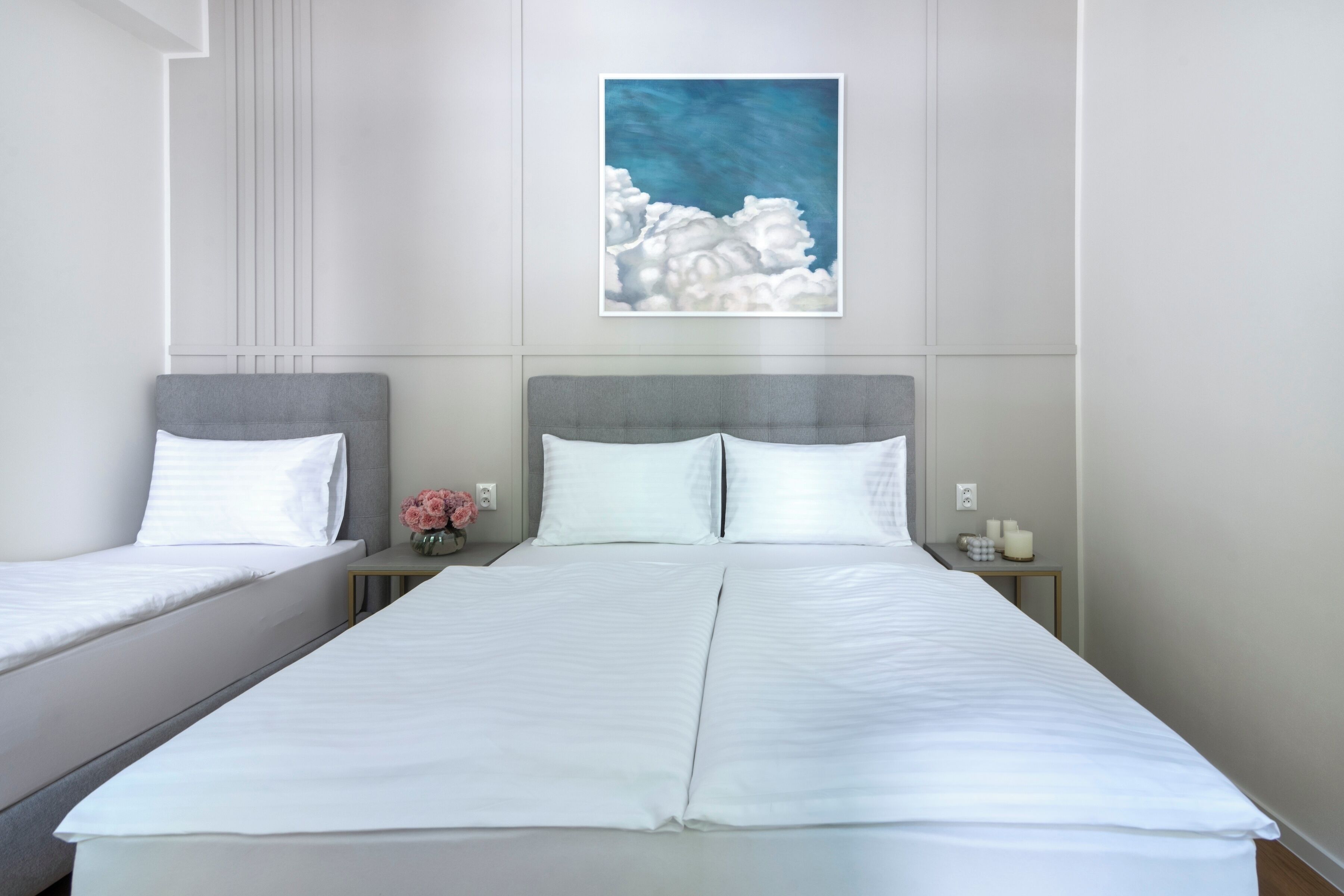 A hotel room with two beds, a large painting of clouds, and a nightstand with a vase of flowers.