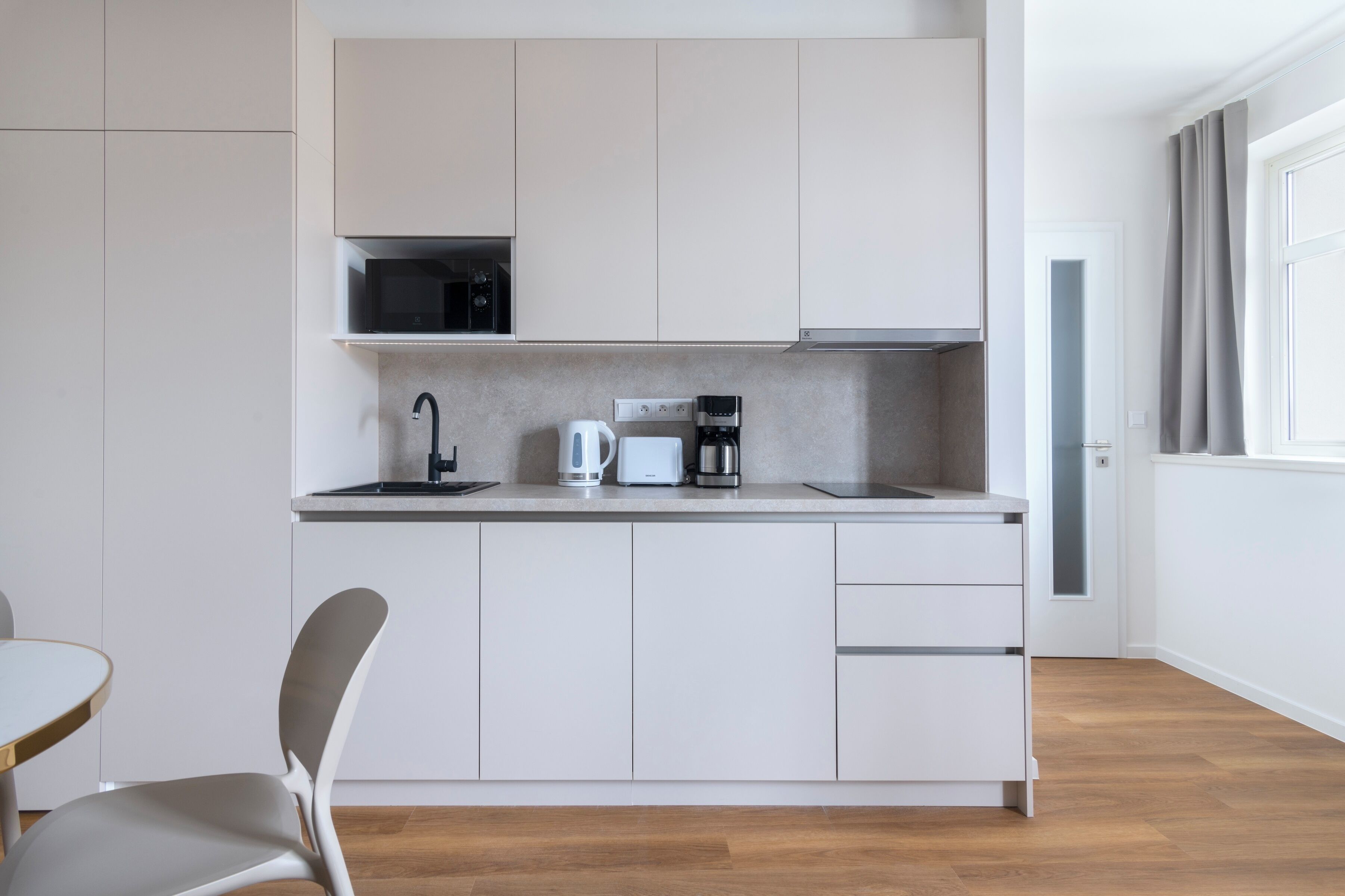 Una cucina moderna con mobili bianchi, un forno a microonde, una macchina per il caffè e un lavello. C'è una sedia e una finestra con tende.