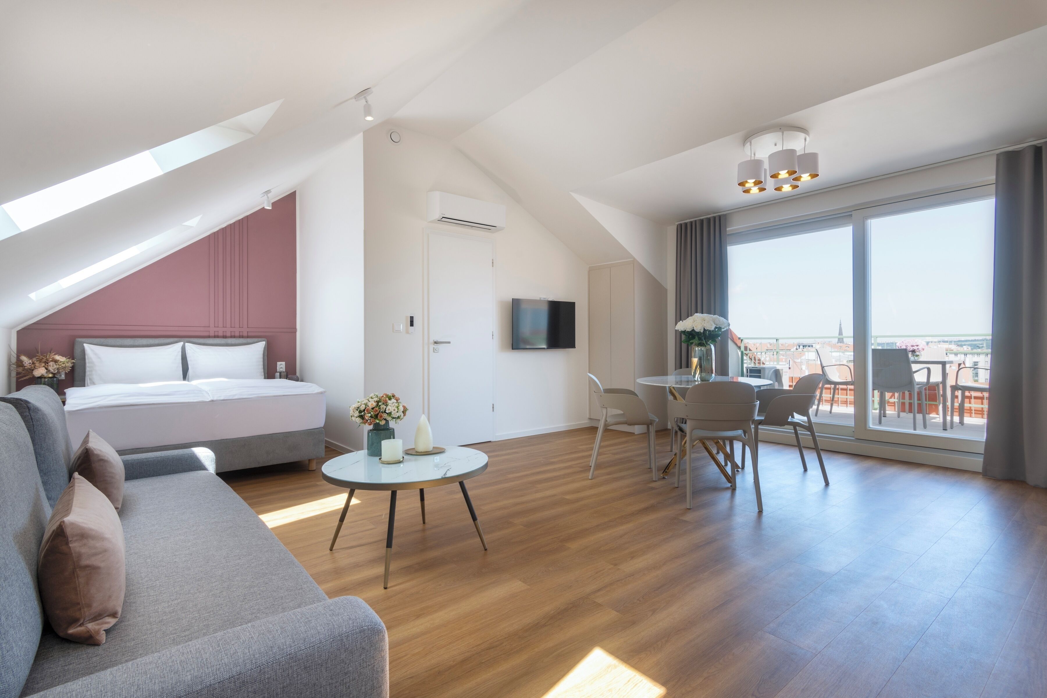 Ein modernes, helles Zimmer mit einem Bett, einer Couch, einem Esstisch mit Stühlen und großen Fenstern mit Stadtblick.