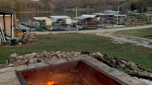 Una chimenea cuadrada con fuego encendido en su interior, rodeada de bloques de piedra. Al fondo, un lago con un muelle y edificios.