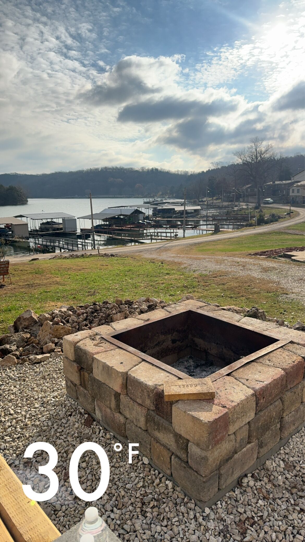 A stone fire pit with a bench, surrounded by gravel, overlooking a marina with docks and boats.