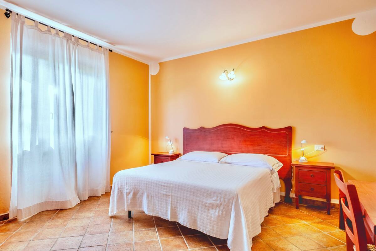 Un dormitorio con cama, dos mesitas de noche, una silla y una ventana con cortinas.