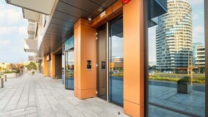 A modern hotel entrance with glass doors, a covered walkway, and a view of a high-rise building in the background. - Stylish Apartment with a Balcony | Air Conditioning (Rzeszów)