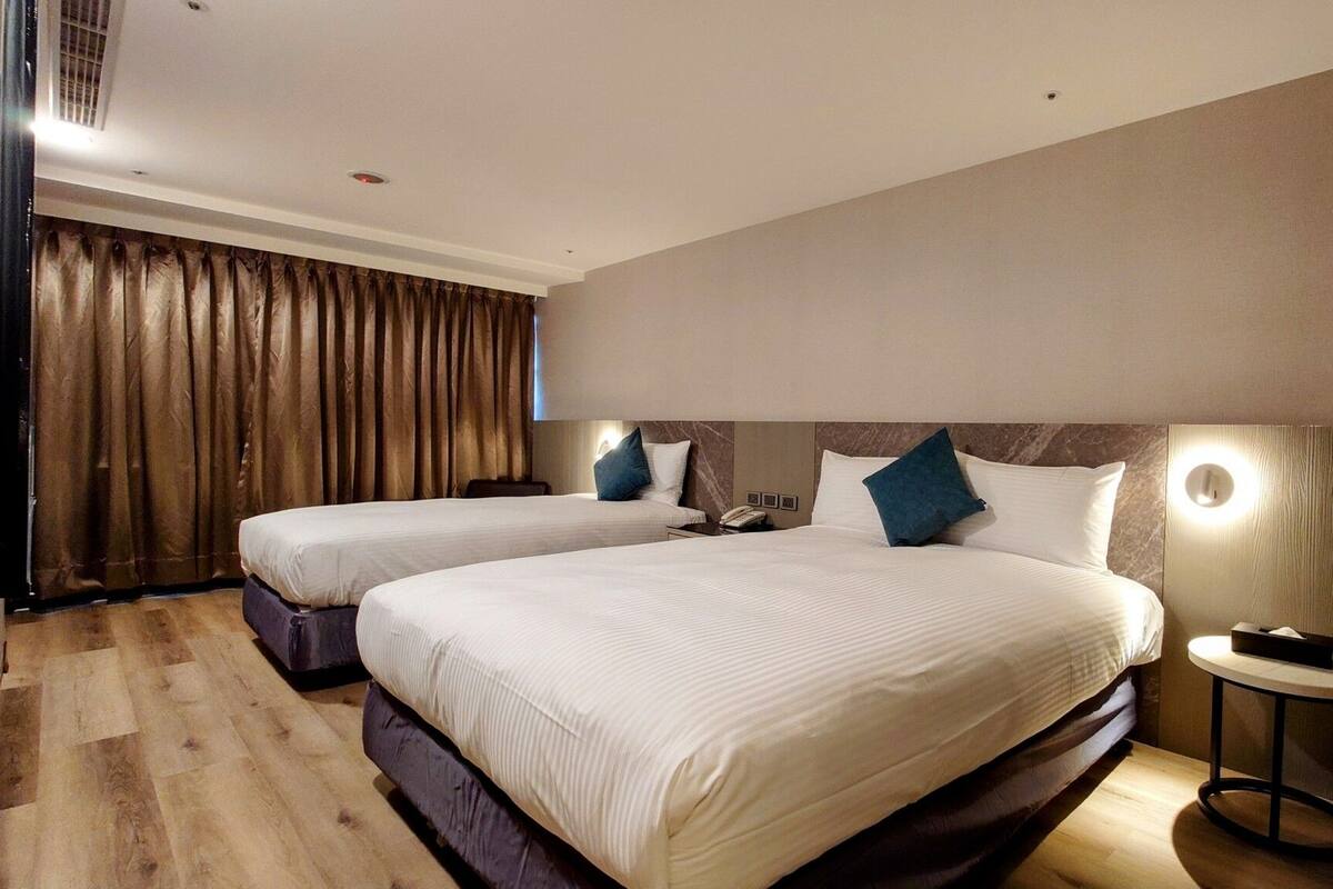 Habitación de hotel con dos camas, suelo de madera, un ventanal grande con cortinas y una mesita de noche con lámpara.