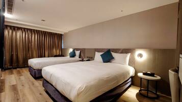 A hotel room with two beds, a wooden floor, a large window with curtains, and a bedside table with a lamp.