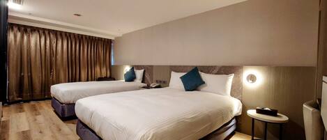 A hotel room with two beds, a wooden floor, a large window with curtains, and a bedside table with a lamp.
