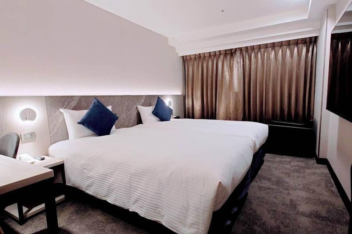 Habitación de hotel con una cama grande, un escritorio y una silla. Hay dos mesitas de noche con lámparas. La habitación tiene una ventana con cortinas.