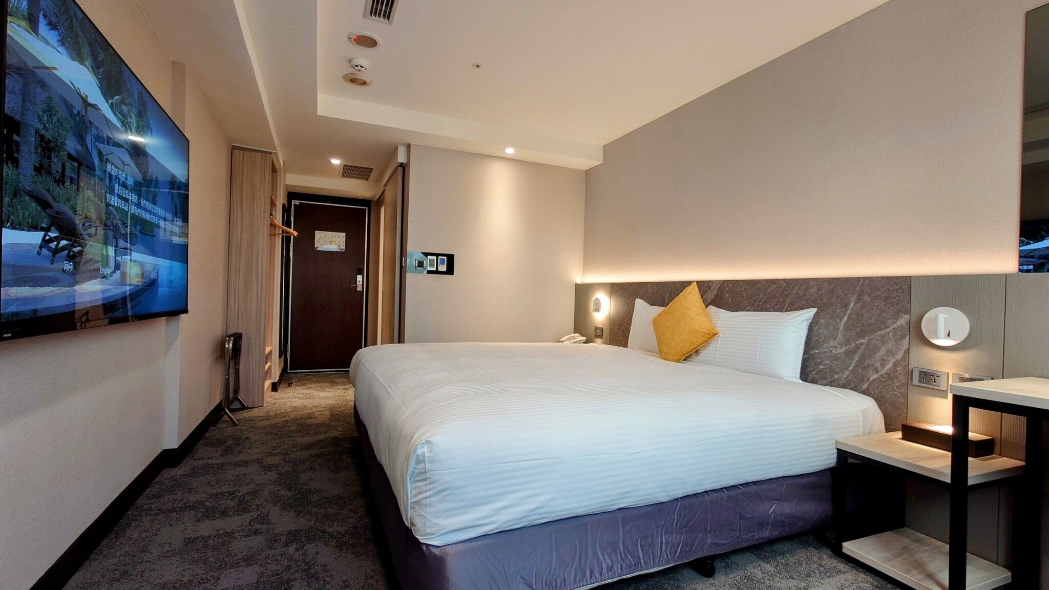 A hotel room with a large bed, a bedside table with a lamp, a wall-mounted TV, and a door leading to the hallway.
