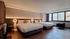 Habitación de hotel con dos camas, un escritorio, una silla y un amplio ventanal con vistas a la ciudad.