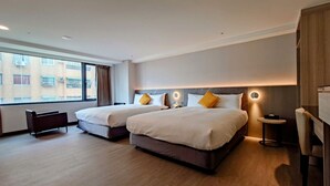 Habitación de hotel con dos camas, un escritorio pequeño y una ventana con vistas a edificios.