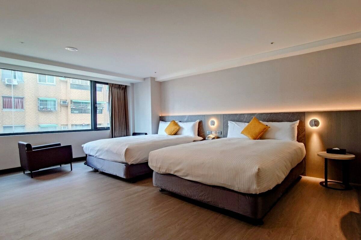 Habitación de hotel con dos camas, un escritorio pequeño y una ventana con vistas a edificios.