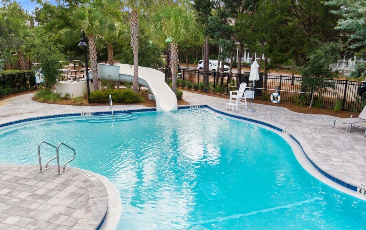Una piscina con tobogán, rodeada de palmeras y un área vallada con sillas de descanso.