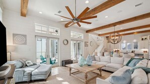 A spacious living room with a large ceiling fan, a staircase, and a kitchen area. - Next to Camp WaterColor! Sleeps 22! Pool, Gym, LSV, Bikes & Carriage House! (Santa Rosa Beach)
