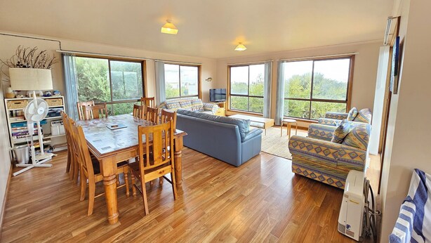 Un vaste espace de vie comprenant une table à manger en bois, un canapé bleu et des fauteuils à motifs. De grandes fenêtres offrent une vue sur la verdure environnante.
