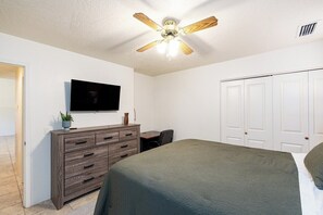 A bedroom with a bed, a dresser, a desk, a ceiling fan, and a TV on the wall.