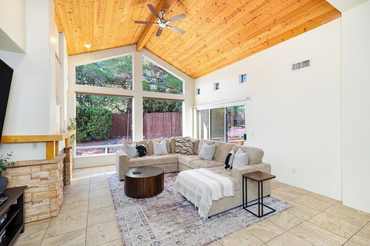 A spacious living room with a wooden ceiling, large windows, a sectional sofa, a coffee table, and a TV.