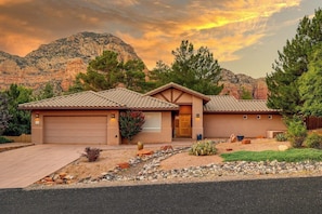 A two-story house with a garage, surrounded by a landscaped garden with rocks and plants, set against a mountainous backdrop.