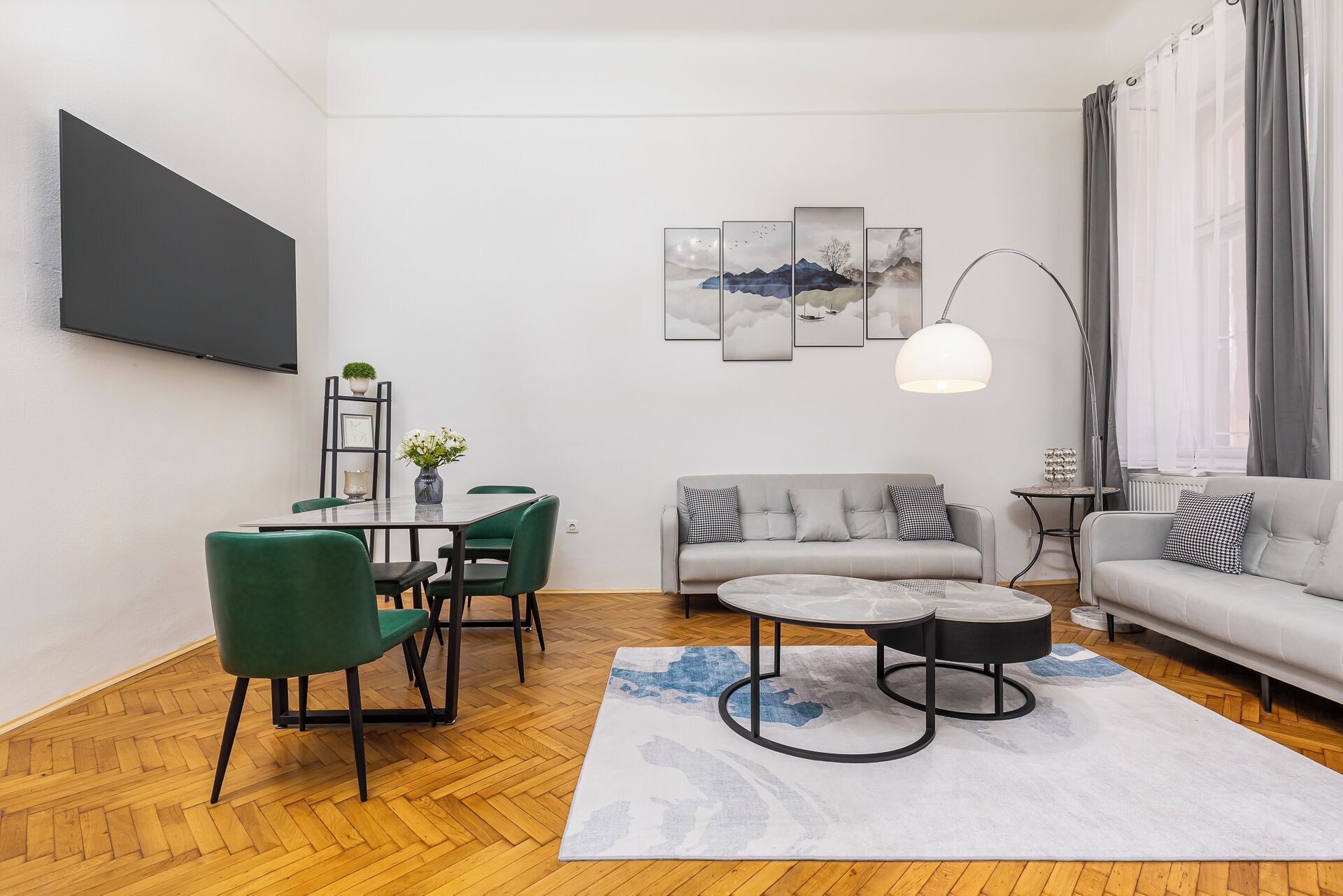 A modern living room with a sofa, a dining table with chairs, a coffee table, a TV mounted on the wall, and a large window with curtains.