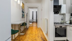 Un couloir étroit doté d’un parquet en bois disposé en chevrons, une petite cuisine sur la droite avec une cuisinière à gaz et un four, et un coin repas avec une chaise verte et une table blanche sur la gauche.