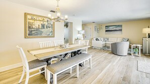 Un comedor con una mesa de madera, sillas blancas y un banco. Sobre la mesa, hay una araña.