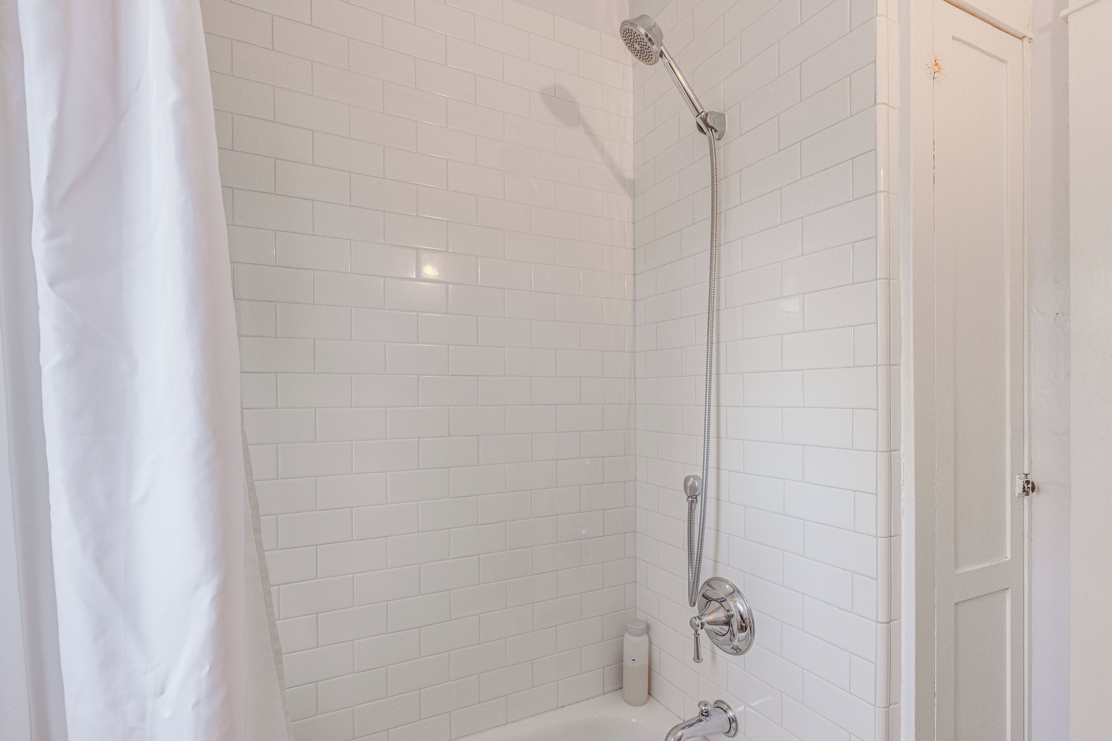 A bathroom with white tiled walls, a shower with a handheld showerhead, and a door.