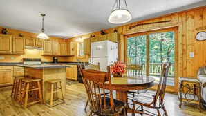 A wooden cabin kitchen with a dining area, wooden bar stools, a refrigerator, and a large window with a view of trees. - Peaceful Wautoma Retreat w/ Deck on 10 Acres! (Wautoma)