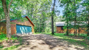 A log cabin with a garage, surrounded by dense forest. - Peaceful Wautoma Retreat w/ Deck on 10 Acres! (Wautoma)