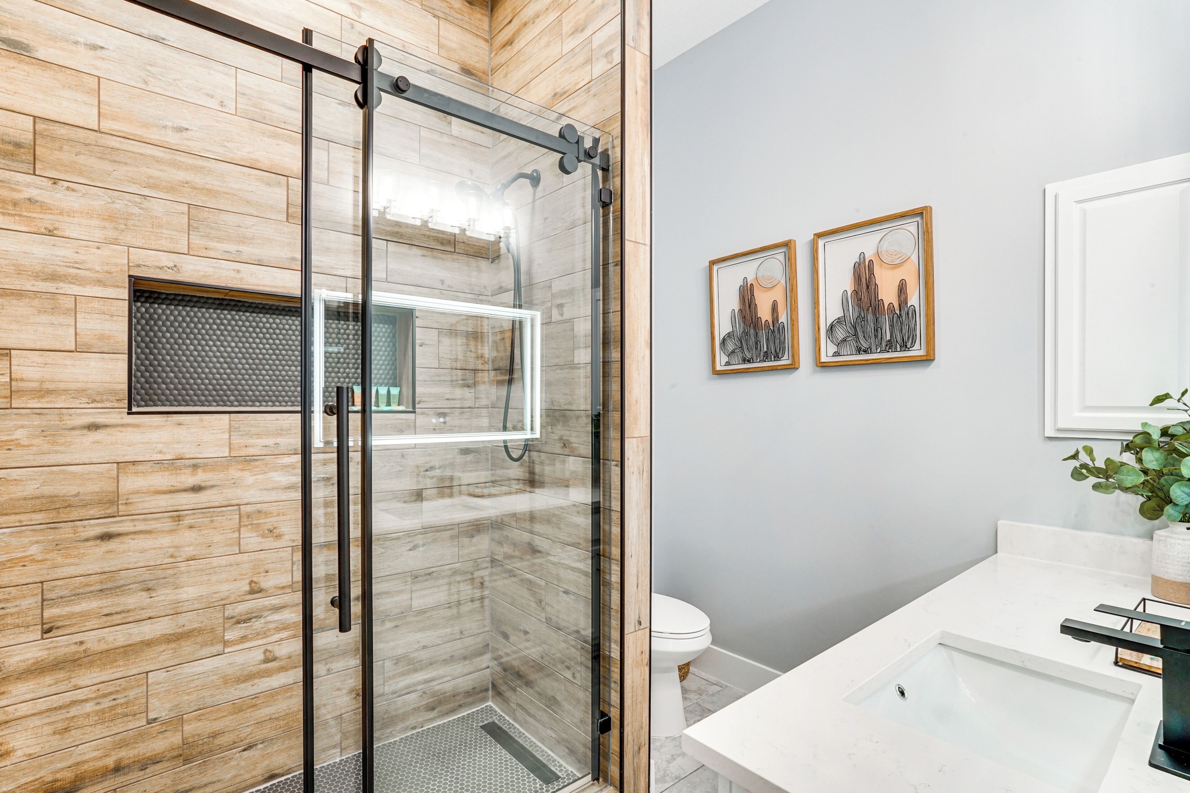 A modern bathroom with a wooden wall, a glass shower enclosure, a white sink, and two framed artworks.