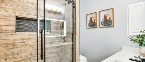 A modern bathroom with a wooden wall, a glass shower enclosure, a white sink, and two framed artworks.