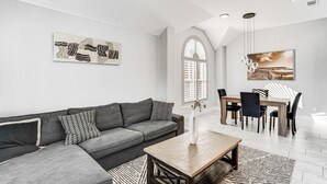Una sala moderna con un sofá seccional gris, una mesa de centro de madera, un comedor con mesa y sillas de madera, y obras de arte en las paredes.