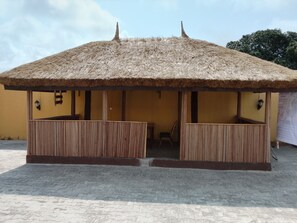 A thatched-roof structure with a wooden floor and walls, featuring two seating areas and a small table. - Palace Holiday Inn LTD (Lekki)