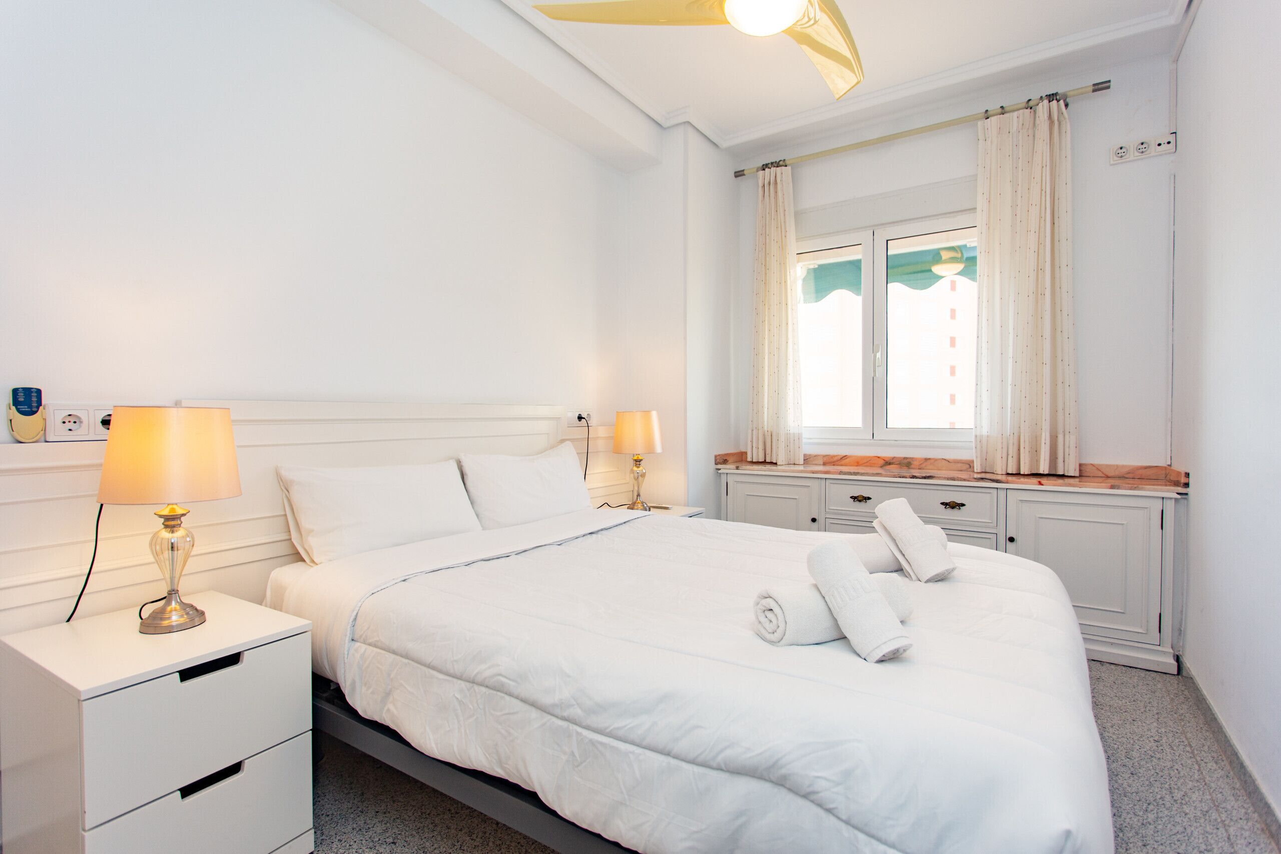 Une chambre à coucher bien rangée, avec un lit, deux tables de chevet dotées de lampes, une fenêtre avec des rideaux et une petite étagère ornée d’objets décoratifs.