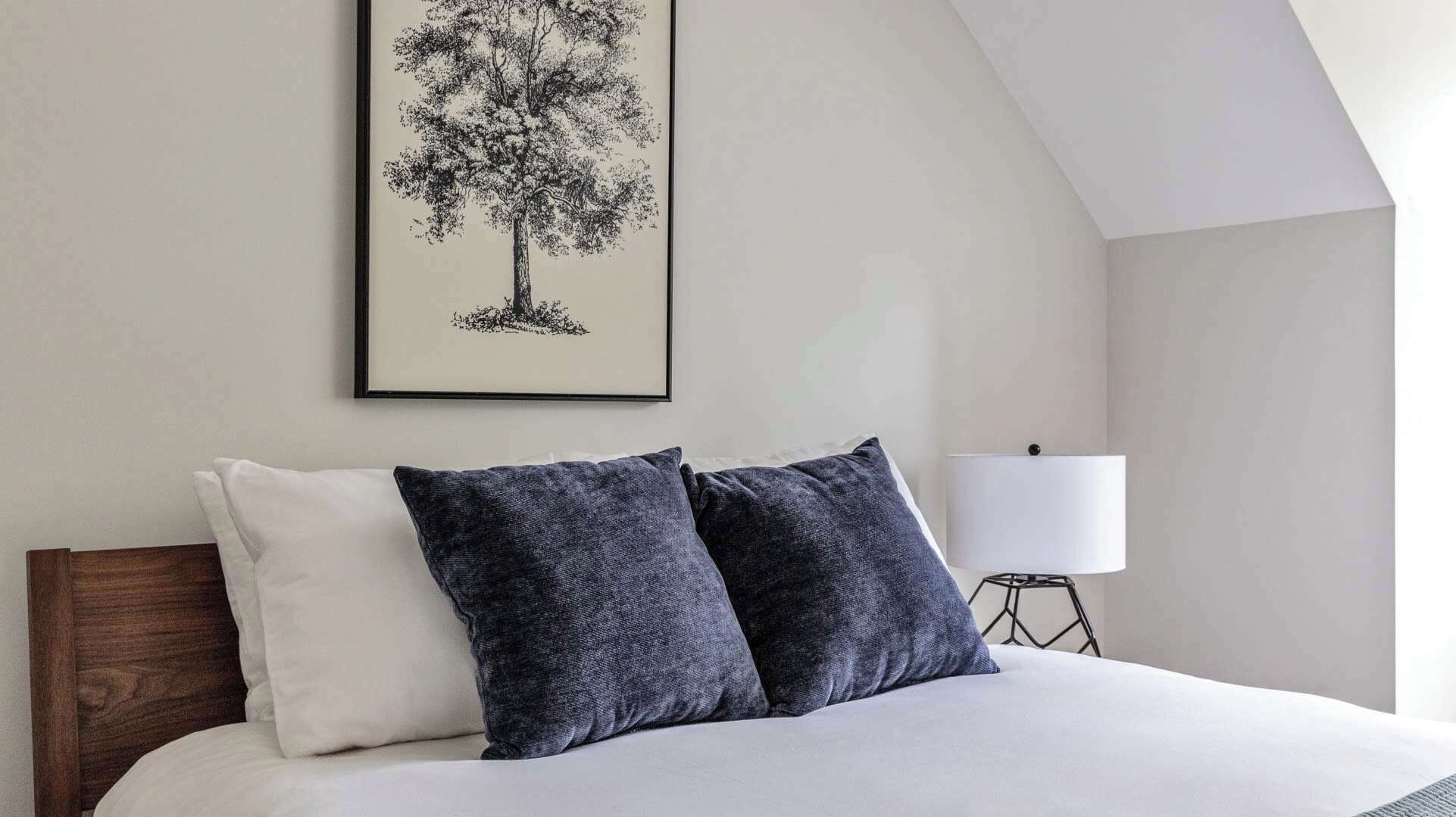 Ein Schlafzimmer mit einem Bett, zwei Kissen, einem gerahmten Baum-Kunstwerk, einer Nachttischlampe und einer geneigten Decke.