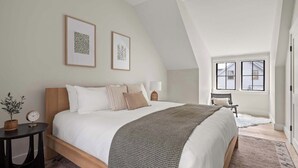 A bedroom with a large bed, two framed pictures on the wall, a bedside table with a lamp, a small potted plant, and a view of the outside through a window.
