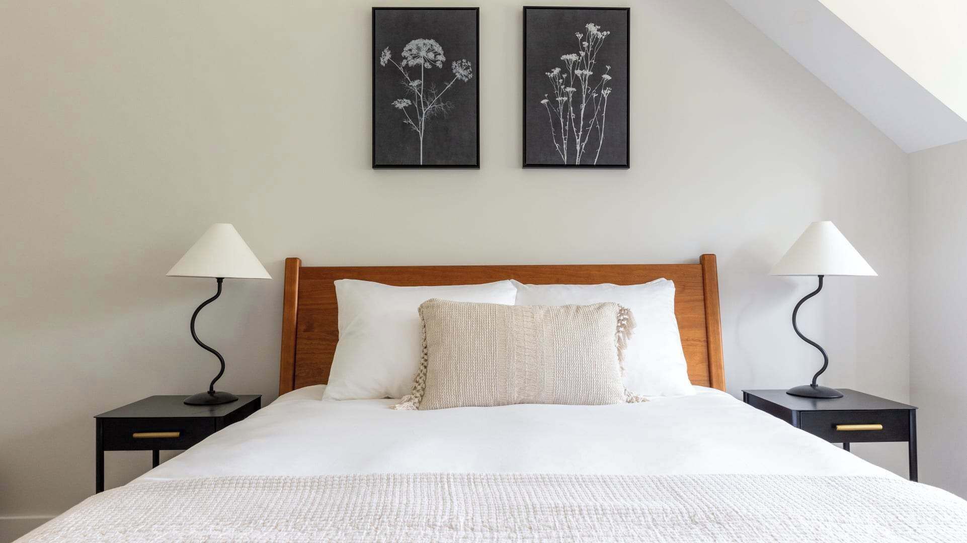 A neatly made bed with white linens, two bedside tables with lamps, and two framed botanical prints on the wall.