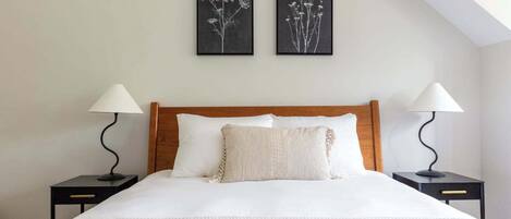 A neatly made bed with white linens, two bedside tables with lamps, and two framed botanical prints on the wall.