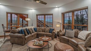 A spacious living room with a large sectional sofa, a coffee table, and a fireplace. There are large windows offering a view of the outdoors. - Extraordinary Location For Large Groups | Apex Grande (Steamboat Springs)
